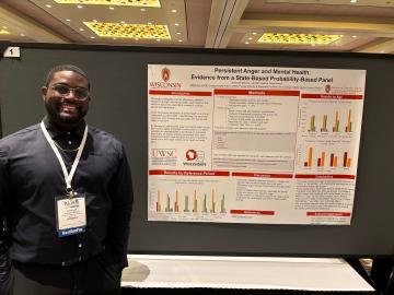 Jeremyah Williams smiles next to a poster titled "Persistent Anger and Mental Health: Evidence from a State-Based Probability-Based Panel"