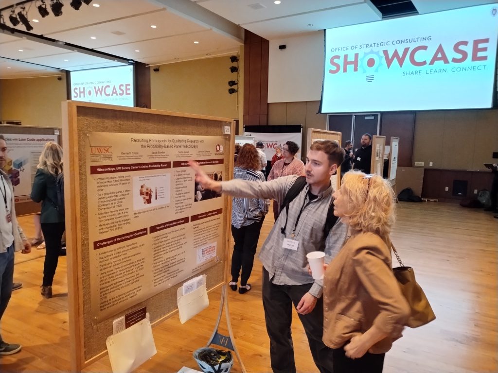 Jacob Boelter stands in front of a board with a blonde woman, pointing out a poster titled Recruiting Participants to Qualitative Research for Probably-Based Panel Wisconsays